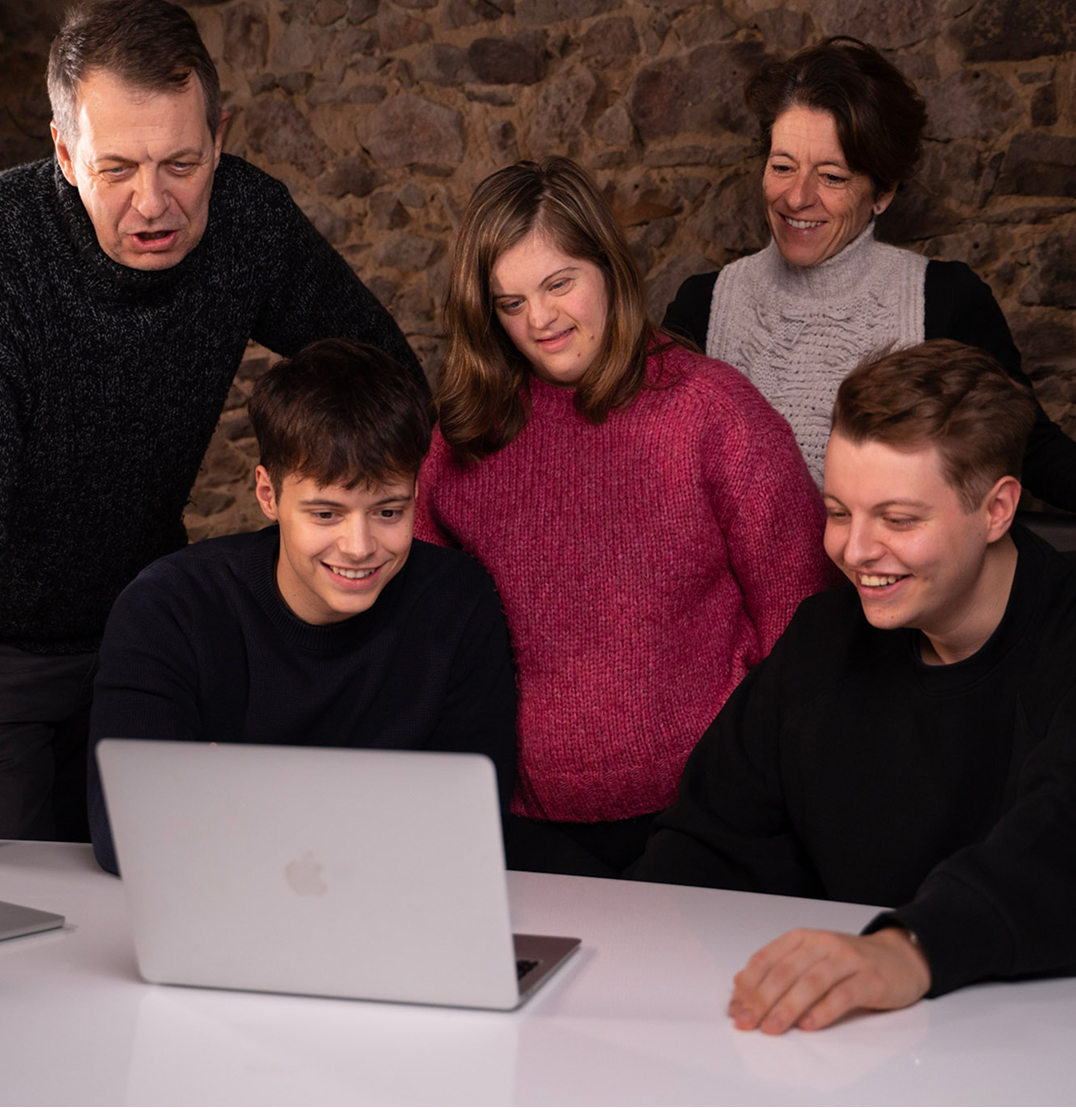 Foto von Familie Merz. Fünf Personen schauen gemeinsam lächelnd auf einen Laptop-Bildschirm.