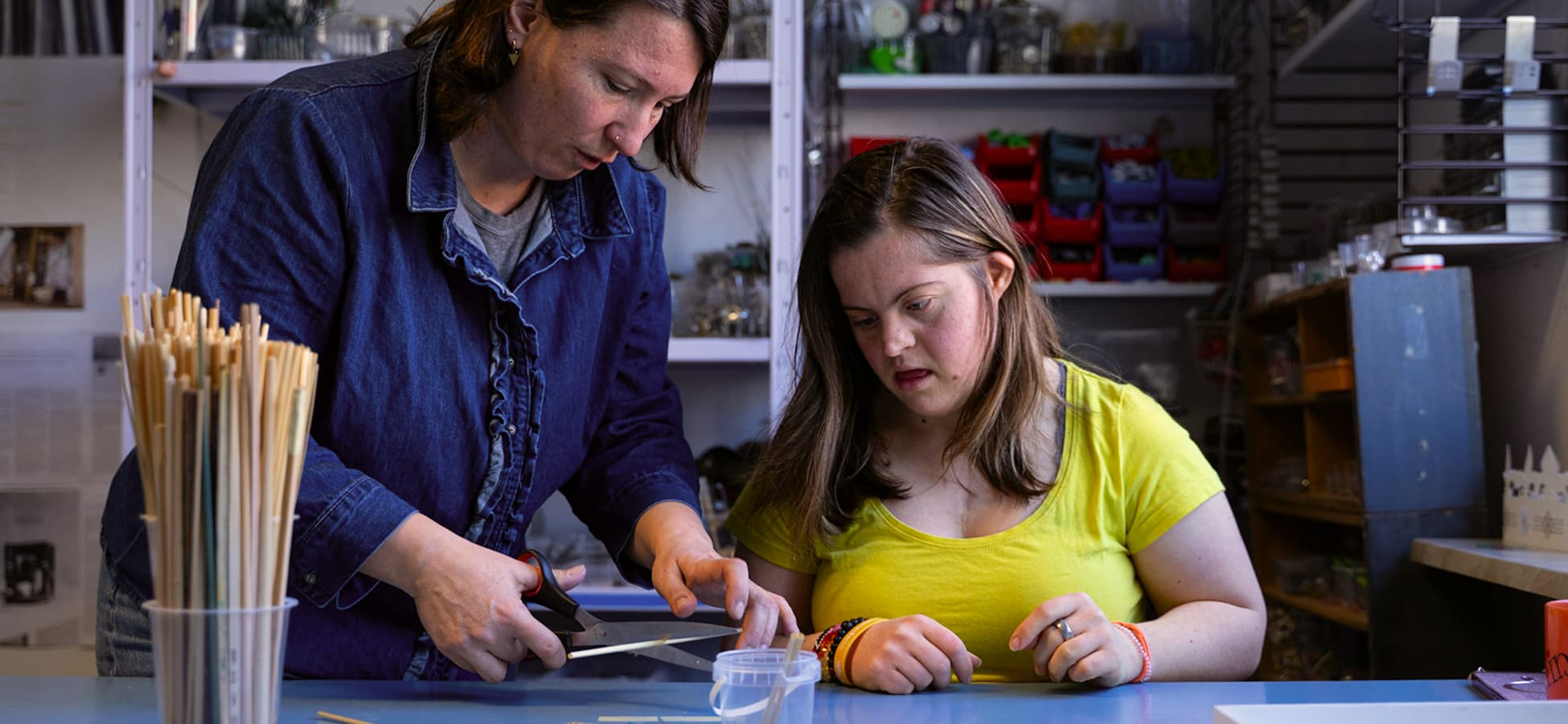 Betreuerin und junge Frau mit gelbem T-Shirt arbeiten gemeinsam mit Schere an einem Werktisch