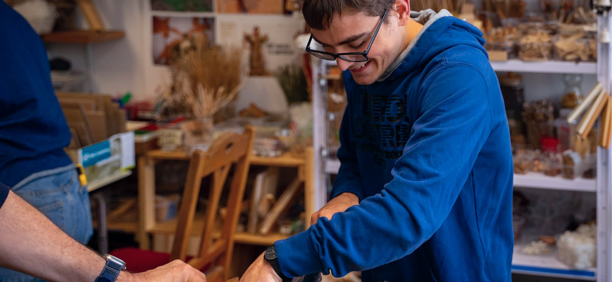 Junger Mann mit Brille und blauem Hoodie arbeitet lächelnd in einer Holz-Werkstatt.