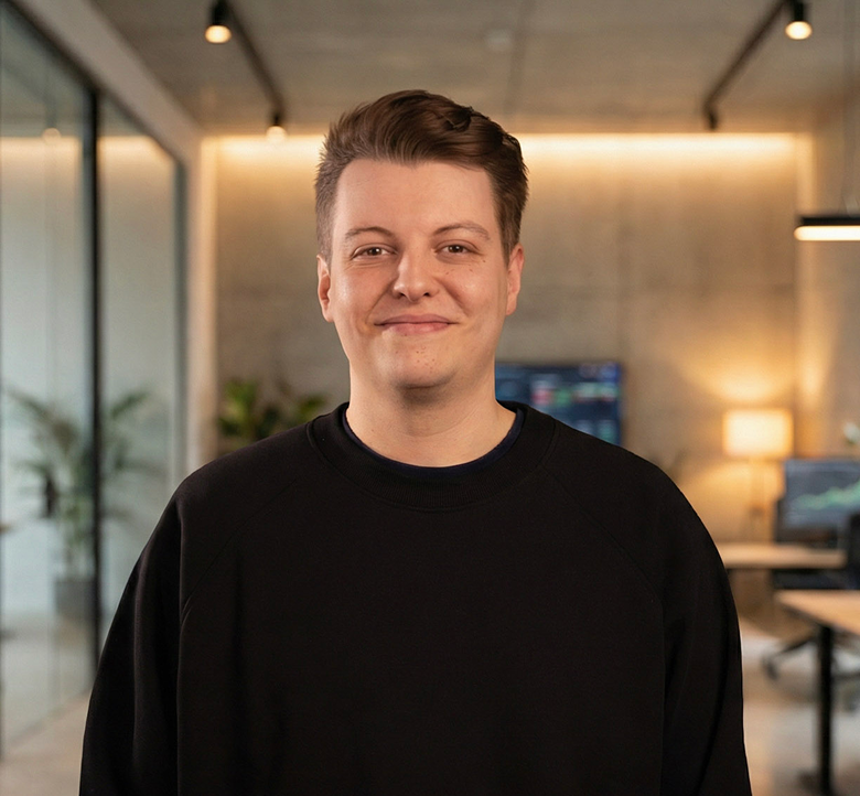 Porträtfoto von Julian Merz – junger Mann in schwarzem Pullover lächelt in einem modernen Büro.